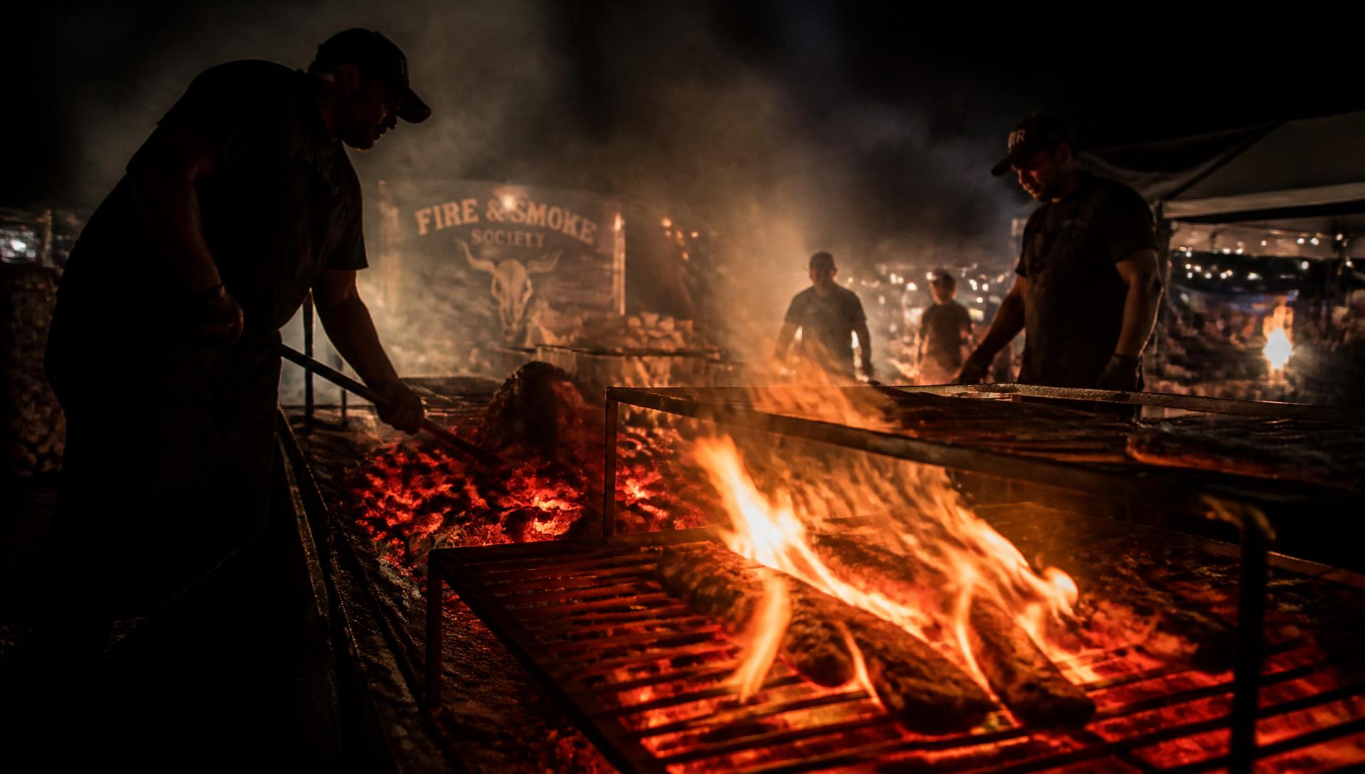 Pit masters working an open fire grill at night, smoke and embers rising